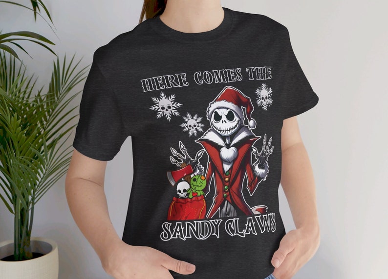 Pu&ograve; includere: Una maglietta nera con una grafica bianca di Jack Skellington vestito da Babbo Natale. La grafica dice "Here Comes The Sandy Claws".