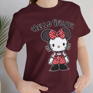 May include: Burgundy t-shirt with a cartoon image of Hello Kitty wearing a red and white polka dot dress and a bow. The text "Hello Disney" is printed on the shirt.