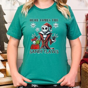 Pu&ograve; includere: Una maglietta verde con un'immagine di Jack Skellington de L'Incubo Prima di Natale vestito da Babbo Natale. Il testo "Here Comes The Sandy Claws" &egrave; stampato sulla maglietta.