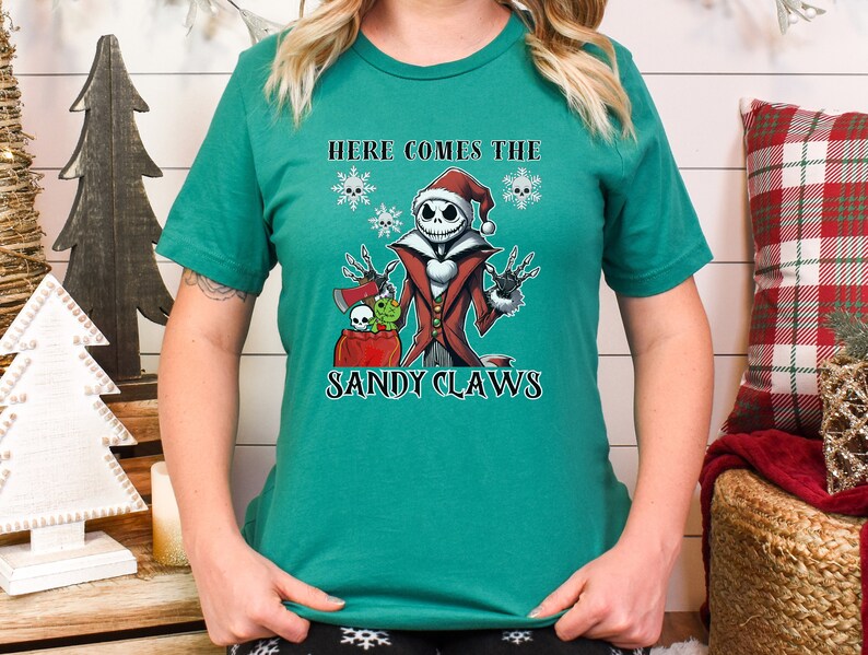 Pu&ograve; includere: Una maglietta verde con un'immagine di Jack Skellington de L'Incubo Prima di Natale vestito da Babbo Natale. Il testo "Here Comes The Sandy Claws" &egrave; stampato sulla maglietta.