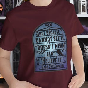 Pode incluir: Camiseta vinho com um gr&aacute;fico azul e branco de uma l&aacute;pide com o texto "Just because cannot see it, doesn't mean I can't believe it!" e "Jack Skellington".