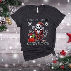 Pu&ograve; includere: Una maglietta grigio scuro con una grafica di Jack Skellington vestito da Babbo Natale. Il testo "Here Comes The Sandy Claws" &egrave; stampato sotto la grafica.