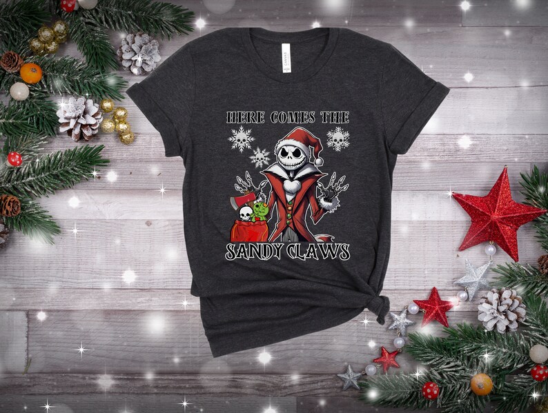 Pu&ograve; includere: Una maglietta grigio scuro con una grafica di Jack Skellington vestito da Babbo Natale. Il testo "Here Comes The Sandy Claws" &egrave; stampato sotto la grafica.