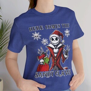 Pu&ograve; includere: Una maglietta blu con un'immagine di Jack Skellington vestito da Babbo Natale. Il testo "Here Comes The Sandy Claws" &egrave; stampato sulla maglietta.