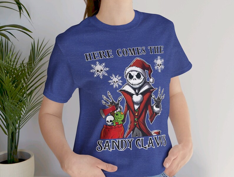 Pu&ograve; includere: Una maglietta blu con un'immagine di Jack Skellington vestito da Babbo Natale. Il testo "Here Comes The Sandy Claws" &egrave; stampato sulla maglietta.
