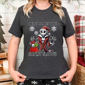 Pu&ograve; includere: Una maglietta grigio scuro con un'immagine di un personaggio dei cartoni animati che indossa un cappello da Babbo Natale e tiene un sacco di regali. Il testo "Here comes the Sandy Claws" &egrave; stampato sotto il personaggio.