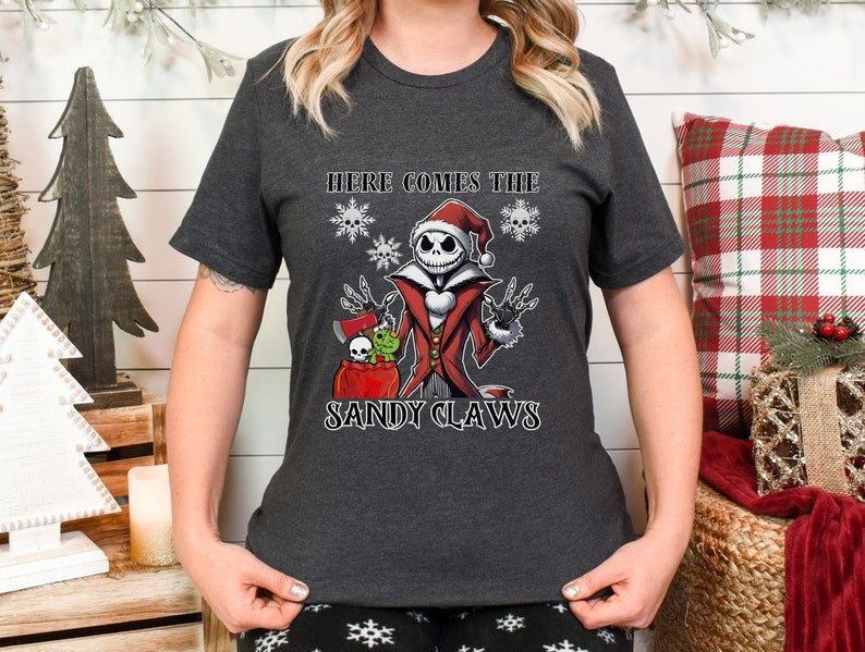 Pu&ograve; includere: Una maglietta grigio scuro con un'immagine di un personaggio dei cartoni animati che indossa un cappello da Babbo Natale e tiene un sacco di regali. Il testo "Here comes the Sandy Claws" &egrave; stampato sotto il personaggio.
