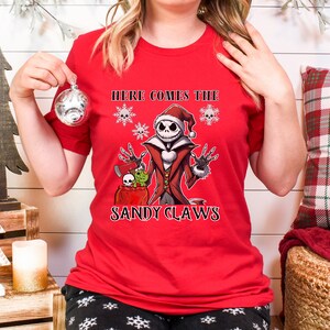Pu&ograve; includere: Maglietta rossa con una grafica di Jack Skellington vestito da Babbo Natale. La grafica include il testo "Here Comes The Sandy Claws".
