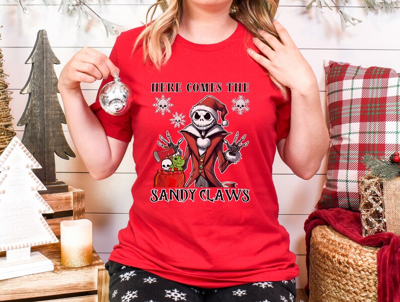 Pu&ograve; includere: Maglietta rossa con una grafica di Jack Skellington vestito da Babbo Natale. La grafica include il testo "Here Comes The Sandy Claws".