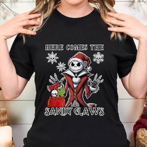 Pu&ograve; includere: Maglietta nera con una grafica di Jack Skellington vestito da Babbo Natale che tiene un sacco di regali. Il testo "Here Comes The Sandy Claws" &egrave; stampato sulla maglietta.