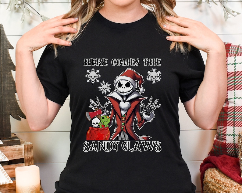 Pu&ograve; includere: Maglietta nera con una grafica di Jack Skellington vestito da Babbo Natale che tiene un sacco di regali. Il testo "Here Comes The Sandy Claws" &egrave; stampato sulla maglietta.