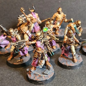 May include: A collection of miniature figurines depicting futuristic warriors. The figures are painted in metallic gold, purple, and brown tones, with detailed weaponry and armor. Each figure is mounted on a textured, circular base, suggesting a tabletop game or collectible.