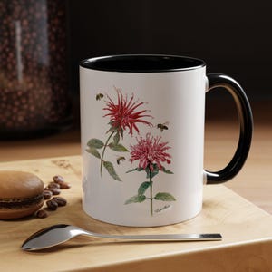 May include: White ceramic mug with a black rim and handle. The mug features a floral design with two pink and red flowers and bees. The text "Lycopus" is printed on the bottom of the mug.