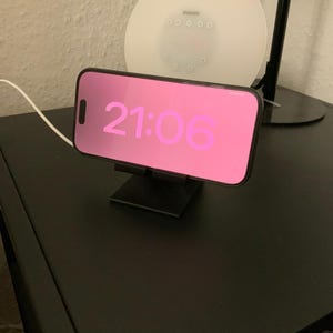 Könnte beinhalten: Ein schwarzes Smartphone mit einem rosa Bildschirm, auf dem die Uhrzeit 21:06 angezeigt wird, liegt auf einem schwarzen Ladeständer.