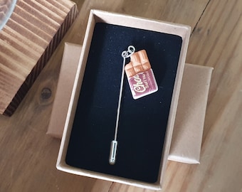 Könnte beinhalten: Silberfarbene Anstecknadel mit einem Schokoladenriegel-Anhänger, präsentiert in einer Geschenkbox. Der Schokoriegel ist braun mit einem rot-weißen Etikett mit der Aufschrift "Choc Soft He". Die Nadel wird auf einer schwarzen Samtoberfläche präsentiert.