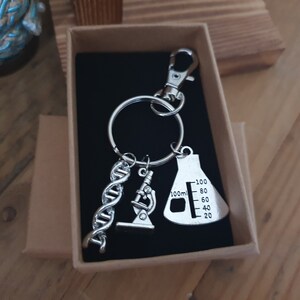 Puede incluir: Llavero plateado con temática científica con un colgante de cadena de ADN, microscopio y matraz. El matraz tiene medidas de hasta 100 ml. Presentado en una pequeña caja de cartón abierta.