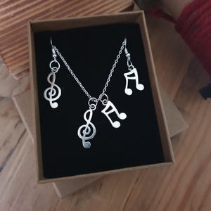 Könnte beinhalten: Silberner Halsketten- und Ohrring-Set mit Musiknoten-Anhängern. Die Halskette hat einen Violinschlüssel-Anhänger und die Ohrringe haben einen Violinschlüssel- und einen Viertelnoten-Anhänger.