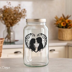 May include: A clear glass jar with a silver lid features a black silhouette of a couple in a heart-shaped design with floral accents. The jar is sitting on a white countertop.