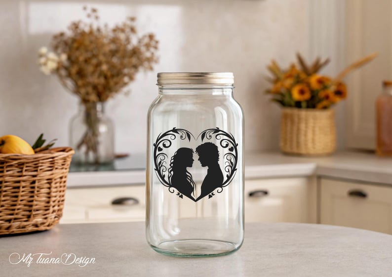 May include: A clear glass jar with a silver lid features a black silhouette of a couple in a heart-shaped design with floral accents. The jar is sitting on a white countertop.