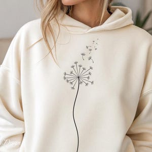 May include: A cream colored hoodie with a black dandelion graphic. The dandelion has seeds blowing in the wind.