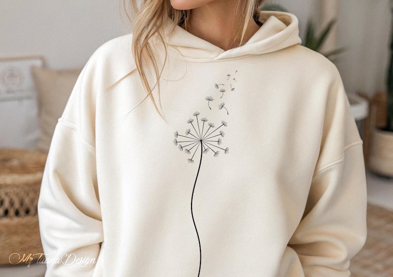 May include: A cream colored hoodie with a black dandelion graphic. The dandelion has seeds blowing in the wind.