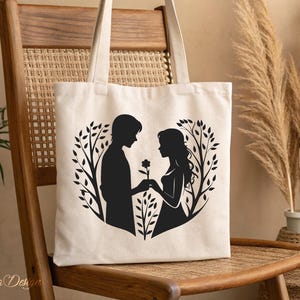 May include: A white canvas tote bag with a black silhouette of a couple holding hands in a heart shape surrounded by branches. The couple is standing in front of a tree with a flower.
