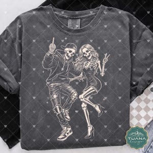 以下が含まれることがあります： チャコールグレーのTシャツで、2体の骸骨が踊っているグラフィックイラストが描かれています。片方の骸骨はパーカー、キャップ、スニーカーを着用し、もう片方はドレスとヒールを着用しています。骸骨は明るいクリーム色で描かれています。