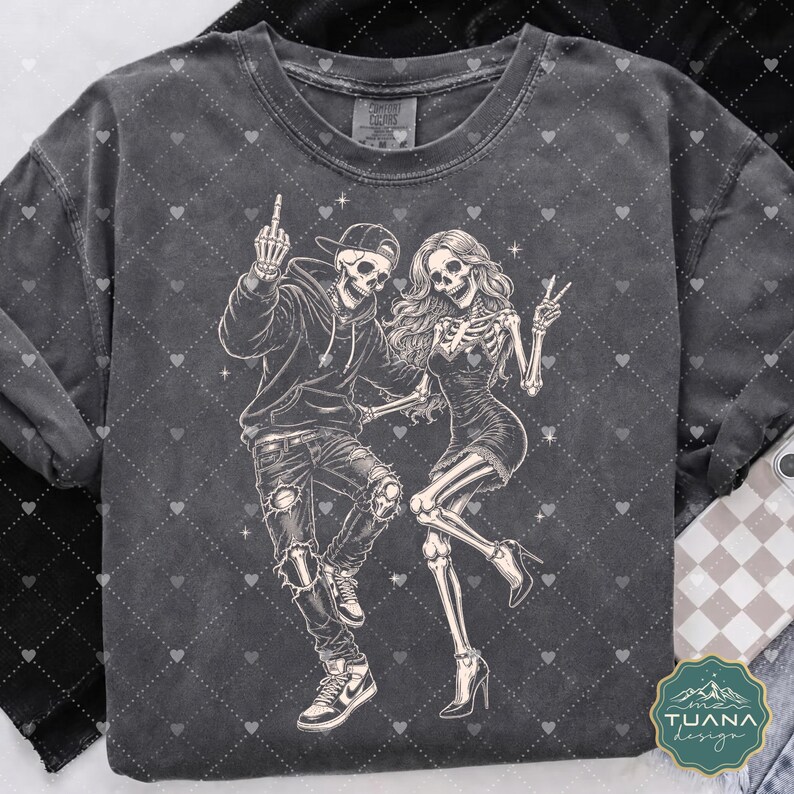 以下が含まれることがあります： チャコールグレーのTシャツで、2体の骸骨が踊っているグラフィックイラストが描かれています。片方の骸骨はパーカー、キャップ、スニーカーを着用し、もう片方はドレスとヒールを着用しています。骸骨は明るいクリーム色で描かれています。