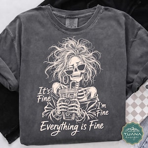 Peut inclure: Un t-shirt gris anthracite à manches courtes avec un motif de squelette aux cheveux ébouriffés, portant des boucles d'oreilles créoles et des lunettes de soleil, tenant une boisson glacée. Le texte indique It's Fine I'm Fine Everything is Fine.