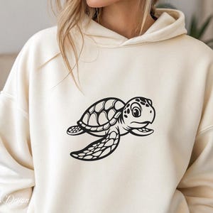 Pu&ograve; includere: Una felpa con cappuccio color crema con una grafica di tartaruga marina nera a cartone animato. La tartaruga nuota verso destra e ha un guscio con un motivo.