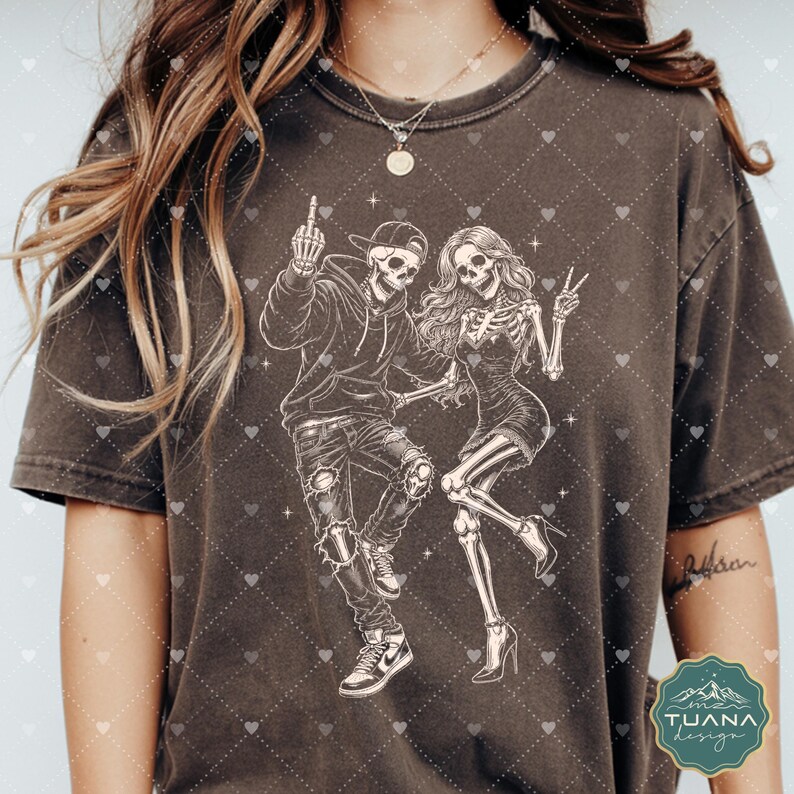 以下が含まれることがあります： ダークチャコールグレーの半袖Tシャツ。2体の骸骨の白い線画が描かれています。1体はパーカー、キャップ、ジーンズを着用し、中指を立てています。もう1体はドレスとハイヒールを着用し、ピースサインをしています。