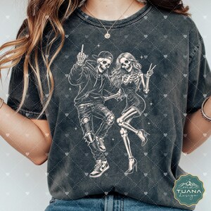 以下が含まれることがあります： チャコールグレーのミネラルウォッシュ加工Tシャツ。2体の骸骨の白いグラフィックイラストが描かれています。1体はパーカー、キャップ、スニーカーを着用し、もう1体はドレスとヒールを着用しています。2体ともダイナミックなダンスのポーズをとっています。