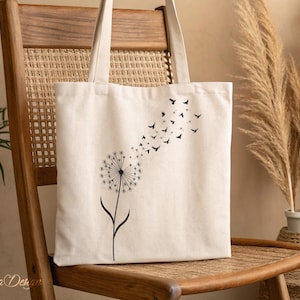 May include: A white canvas tote bag with a black dandelion design and a flock of birds flying away from the dandelion. The bag is sitting on a wooden chair.
