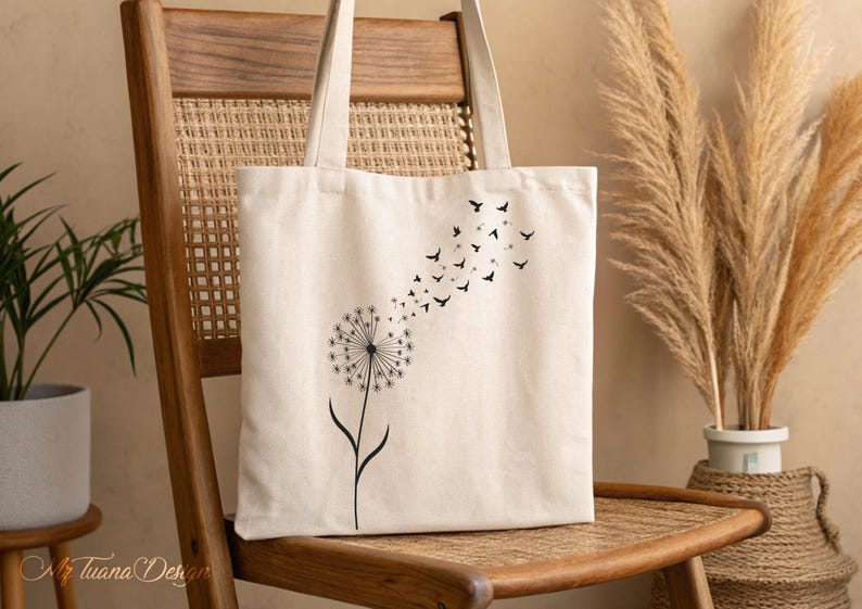 May include: A white canvas tote bag with a black dandelion design and a flock of birds flying away from the dandelion. The bag is sitting on a wooden chair.