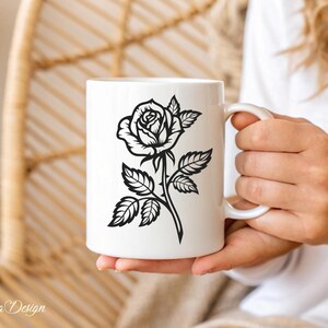 May include: White ceramic mug with a black silhouette of a rose with thorns and leaves. The mug is being held by a hand.