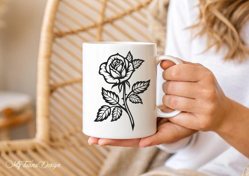 May include: White ceramic mug with a black silhouette of a rose with thorns and leaves. The mug is being held by a hand.