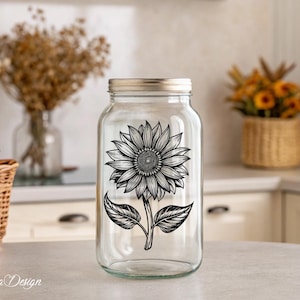 Puede incluir: Un tarro de vidrio transparente con un dise&ntilde;o de girasol en blanco y negro. El tarro tiene una tapa plateada y est&aacute; sentado en una mesa blanca.