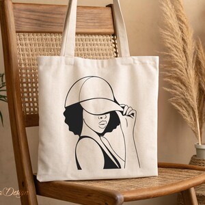 May include: A white tote bag with a black line drawing of a person wearing a baseball cap. The person is looking to the side and has their hand up to their face.