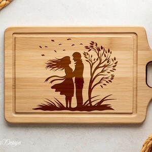 May include: A wooden cutting board with a laser-engraved design of a couple standing under a tree with leaves blowing in the wind. The design is in brown on the natural wood.