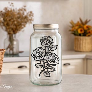 May include: A clear glass jar with a black silhouette of three roses and leaves on the front. The jar has a silver lid.