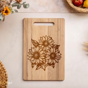 Puede incluir: Una tabla de cortar de madera con un dise&ntilde;o de tres girasoles grabado con l&aacute;ser. Los girasoles son marrones y tienen un dise&ntilde;o detallado.