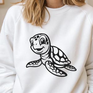 Pu&ograve; includere: Felpa bianca con una grafica di tartaruga marina a cartoni animati in bianco e nero. La tartaruga sorride e ha un guscio con un motivo a pois.