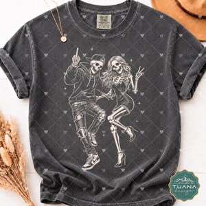 以下が含まれることがあります： チャコールグレーの半袖Tシャツ。2体の骸骨の白いグラフィックプリントが施されています。片方はパーカー、キャップ、スニーカーを着用し、もう片方はドレスとヒールを着用しています。クルーネックで、ヴィンテージ風のウォッシュ加工が施されています。
