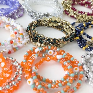 May include: A collection of beaded bracelets in various colors, including orange, white, gold, and purple. Some bracelets feature letter beads spelling out words and phrases. The bracelets are made with small, colorful beads and are designed to be worn as accessories.