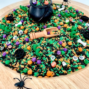 May include: A Halloween-themed sensory bin with black, green, and orange rice. Includes a black cauldron, wooden scoop, plastic spiders, and various Halloween-themed trinkets like pumpkins, skulls, and bones. The bin is set on a wooden tray.