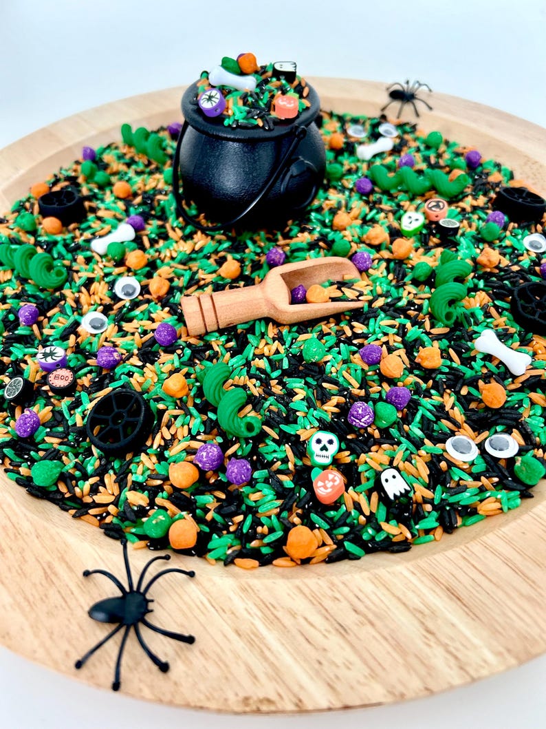 May include: A Halloween-themed sensory bin with black, green, and orange rice. Includes a black cauldron, wooden scoop, plastic spiders, and various Halloween-themed trinkets like pumpkins, skulls, and bones. The bin is set on a wooden tray.