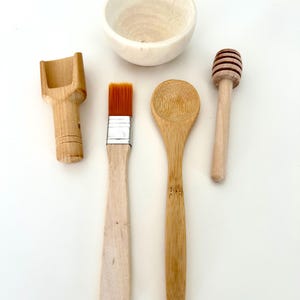 May include: A collection of wooden kitchen utensils and a small bowl. The set includes a scoop, a paintbrush, a bamboo spoon, and a honey dipper. The items are arranged on a white surface, showcasing their natural wood tones.