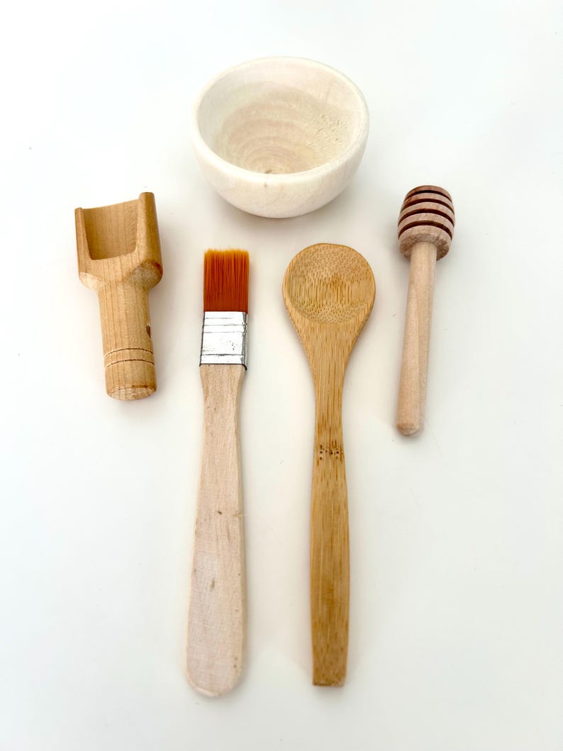 May include: A collection of wooden kitchen utensils and a small bowl. The set includes a scoop, a paintbrush, a bamboo spoon, and a honey dipper. The items are arranged on a white surface, showcasing their natural wood tones.