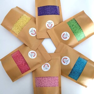 May include: Six brown paper pouches, each containing dyed rice in a different colour: yellow, purple, green, blue, pink, and red. The rice is visible through a clear window. Each pouch has a sticker with a rainbow and the text "Happy Munchkins".
