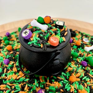 May include: A black cauldron filled with colorful Halloween-themed sensory rice. The cauldron is surrounded by green, orange, black, and purple rice, with small decorative pieces like bones and pumpkins. The cauldron has a black handle.